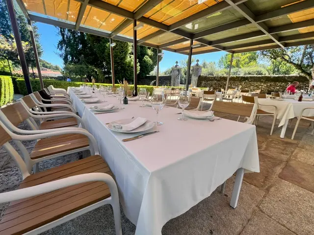 Terraza cubierta con techo de madera y mesas largas con manteles blancos, abierta al jardín
